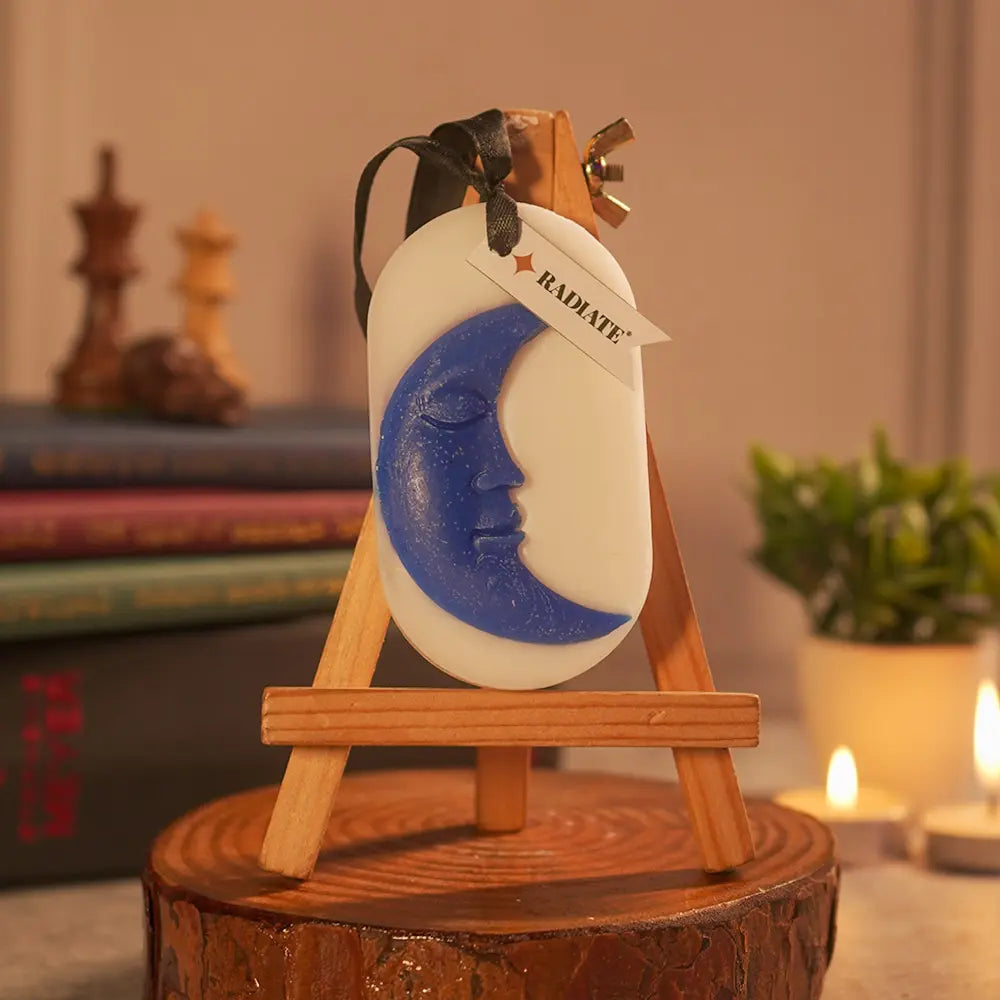 Decorative moon face design embed on a scented wax sachet by Radiate on a wooden stand with a blurred background. Used for sweet scent in closed spaces like almirah, boxes, clothes, shoe rack, gym, tables etc