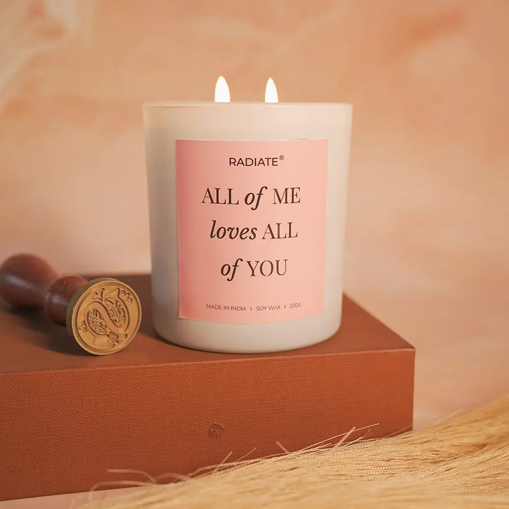 All of me lotus and musk scented candle with a pink label on a wooden block, with a soft peach background